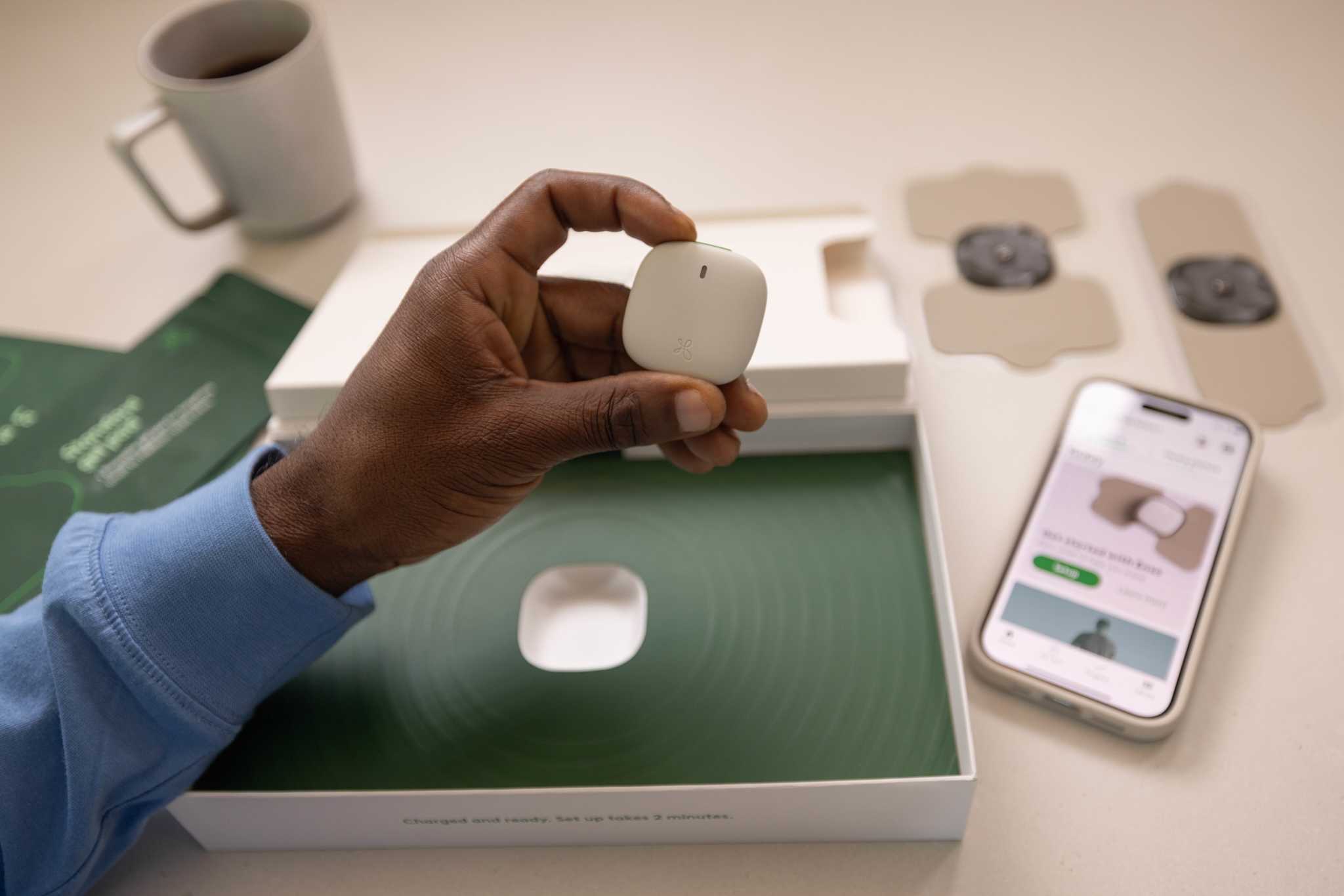Image of the a person's hand holding the Enso 3 device in his fingertips, with the Enso 3 box and gel pads in the background, and a phone nearby with the Hinge Health app displayed
