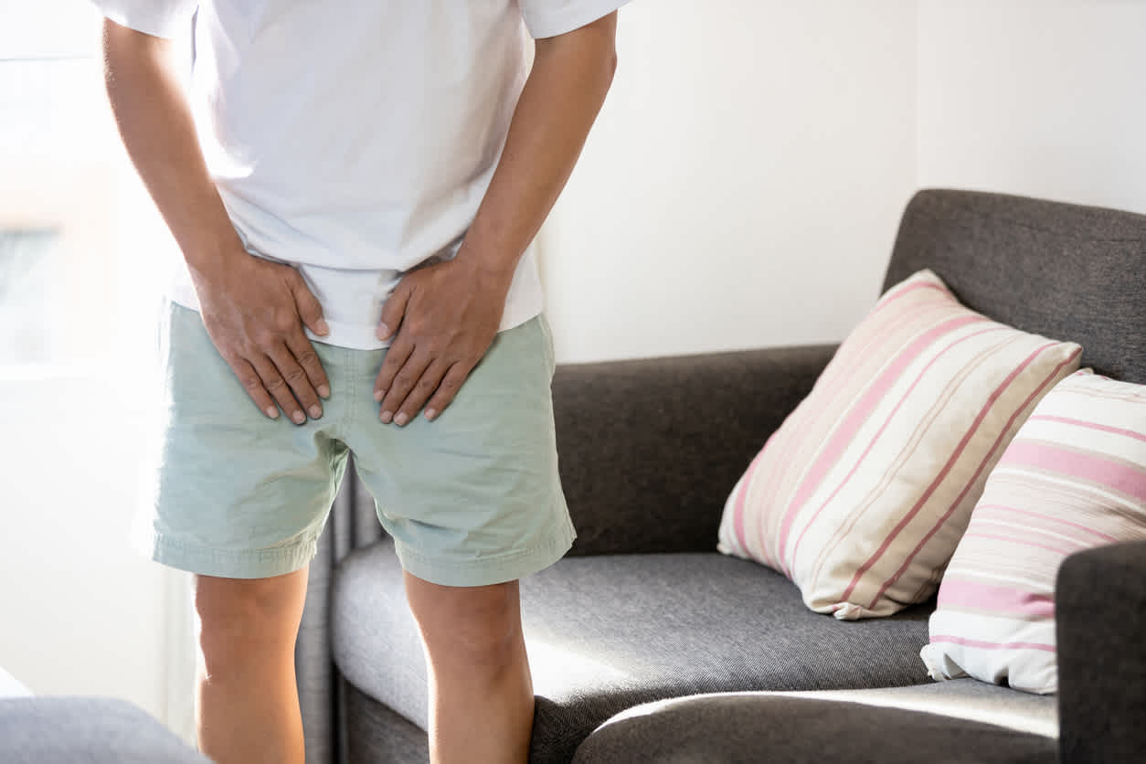 Person in white shirt and mint shorts showing signs of groin pain, standing by gray couch with striped pink pillows