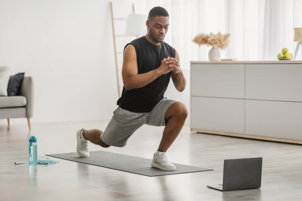 Person doing leg mobility exercises on exercise mat at home, following workout on laptop, with water bottle nearby in bright modern living room