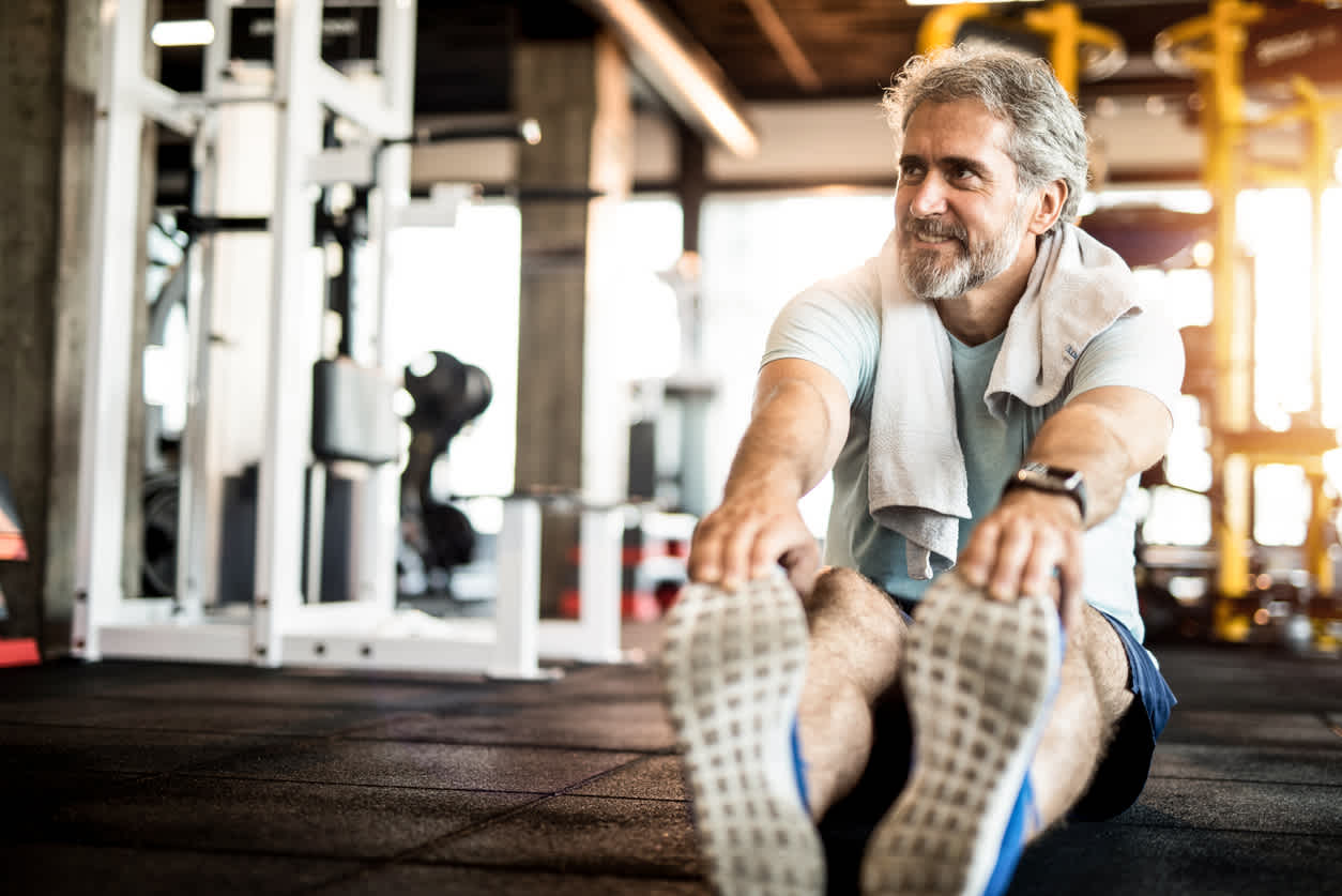 Mature man with gray beard stretching on gym floor, wearing light t-shirt with towel around neck, smiling during workout