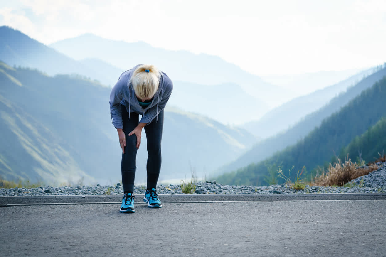 Runner in gray athletic wear and blue shoes pauses to hold knee on mountain road with misty valley view behind