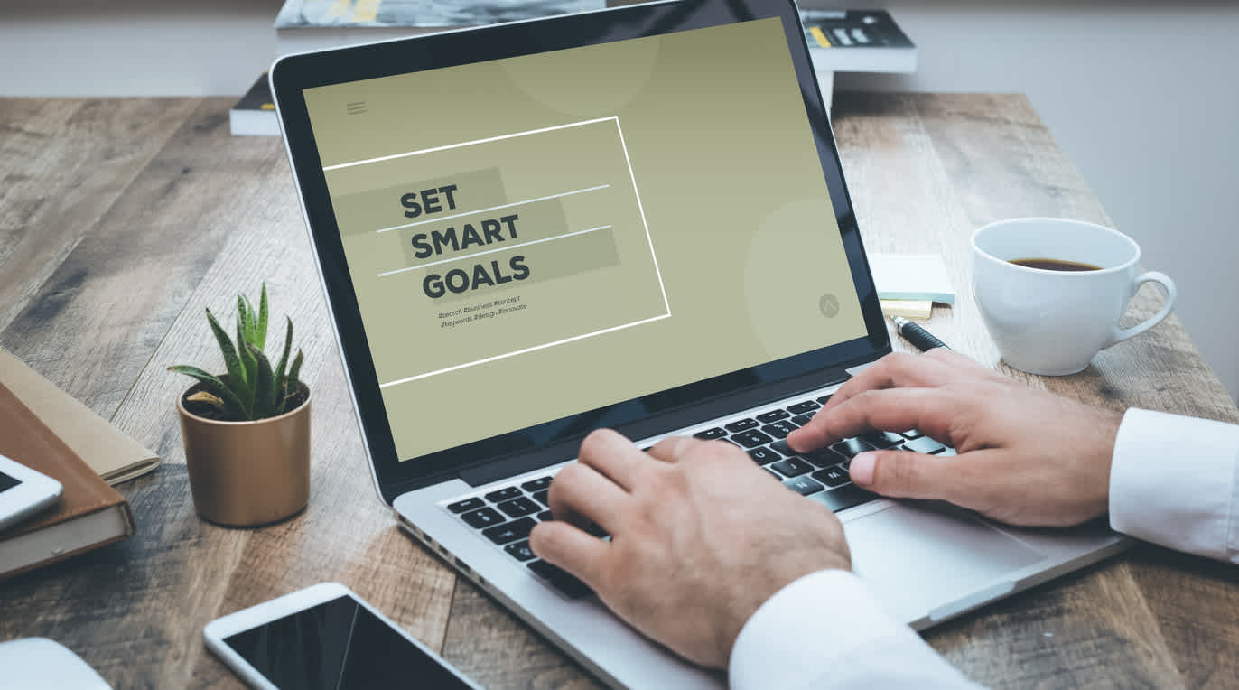 Laptop screen showing "Set Smart Goals" text on olive background, with hands typing, coffee cup and succulent plant on wooden desk