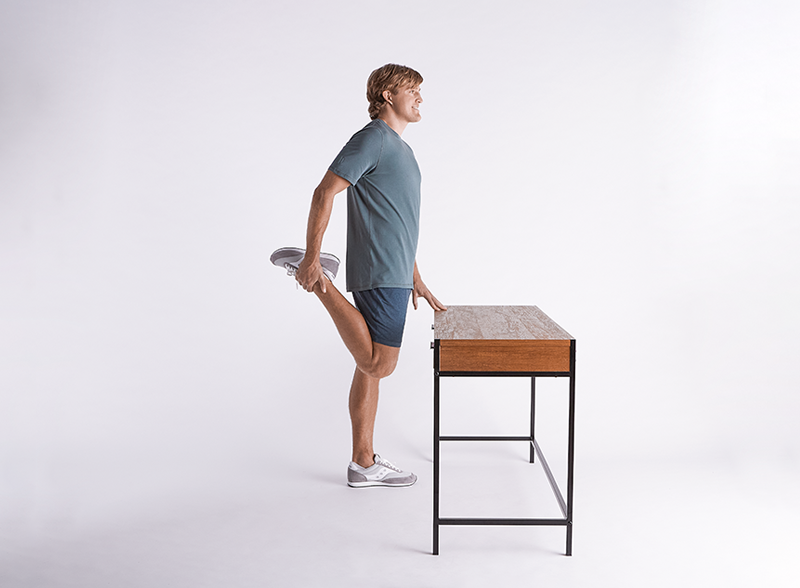 Man-doing-quad-stretch-while-gently-leaning-on-table